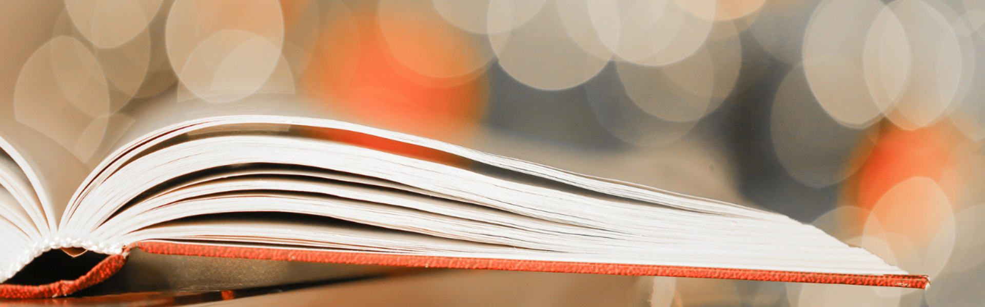 An open book, with an orange cover, lay open on a flat surface with a blurred sparkly background or white, orange and grey orbs.