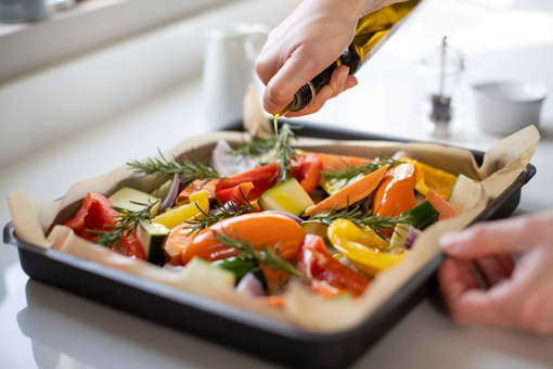 A person drizzling olive oil over a baking tray of Mediterranean vegetables and rosemary herb sprigs