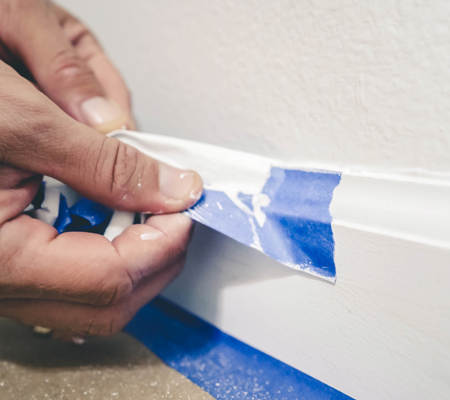 a person peeling back blue tape from a white painting wall and skirting board