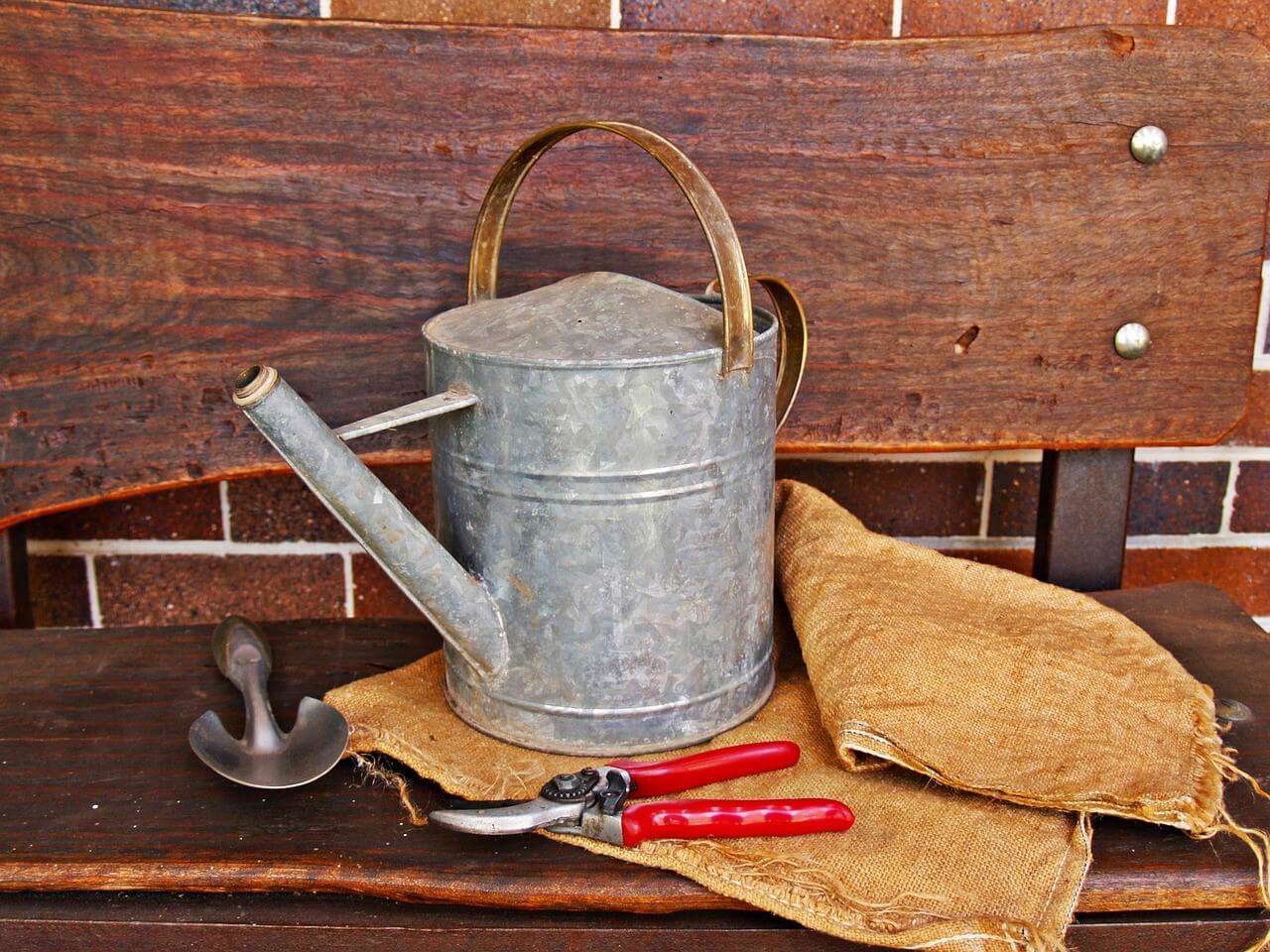 a watering can and red garden sheers and a garden trowel laid out on a wooden table against a brick wall