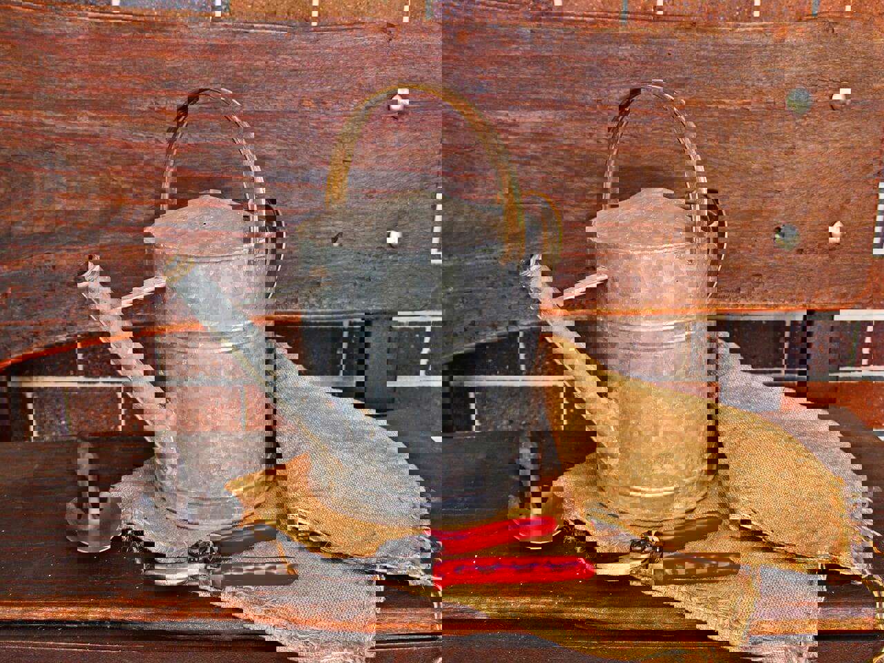 a watering can and red garden sheers and a garden trowel laid out on a wooden table against a brick wall