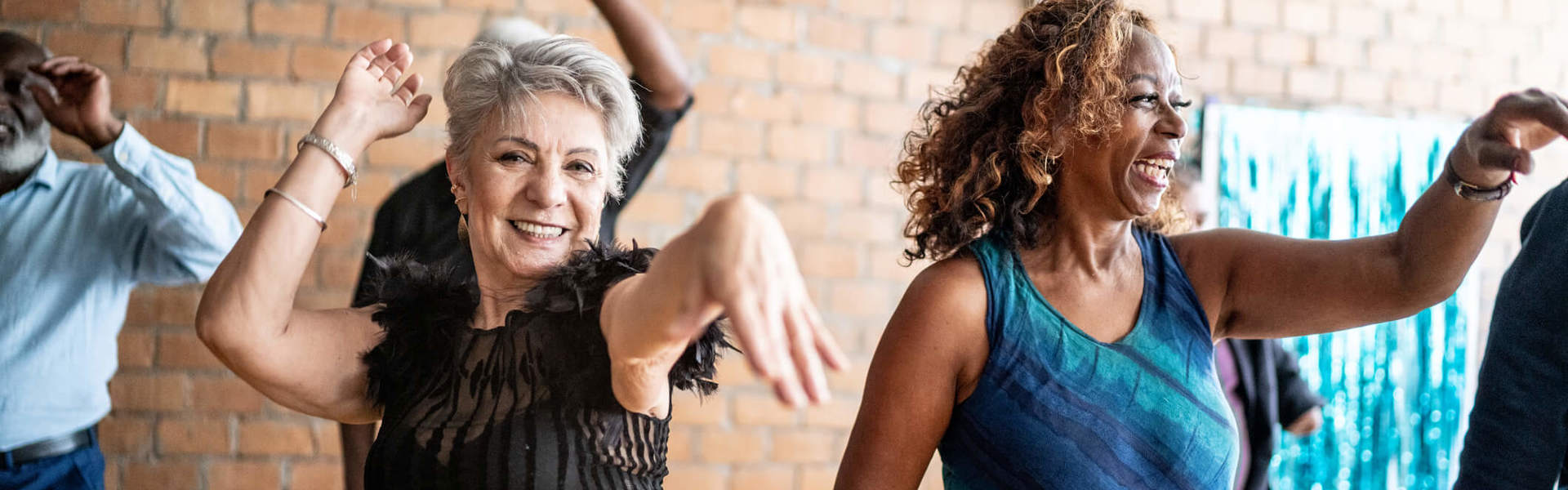 Two older ladies, smiling, laughing and having fun dancing in a group against a brick wall backdrop.