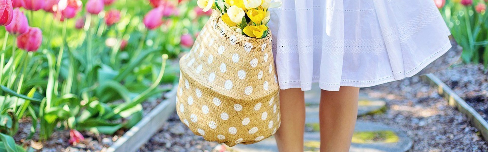 a person wearing a light blue summer dress standing barefoot in the garden holding a polka dot wicker bag filled with yellow flowers