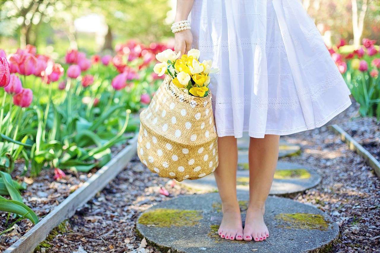 a person wearing a light blue summer dress standing barefoot in the garden holding a polka dot wicker bag filled with yellow flowers