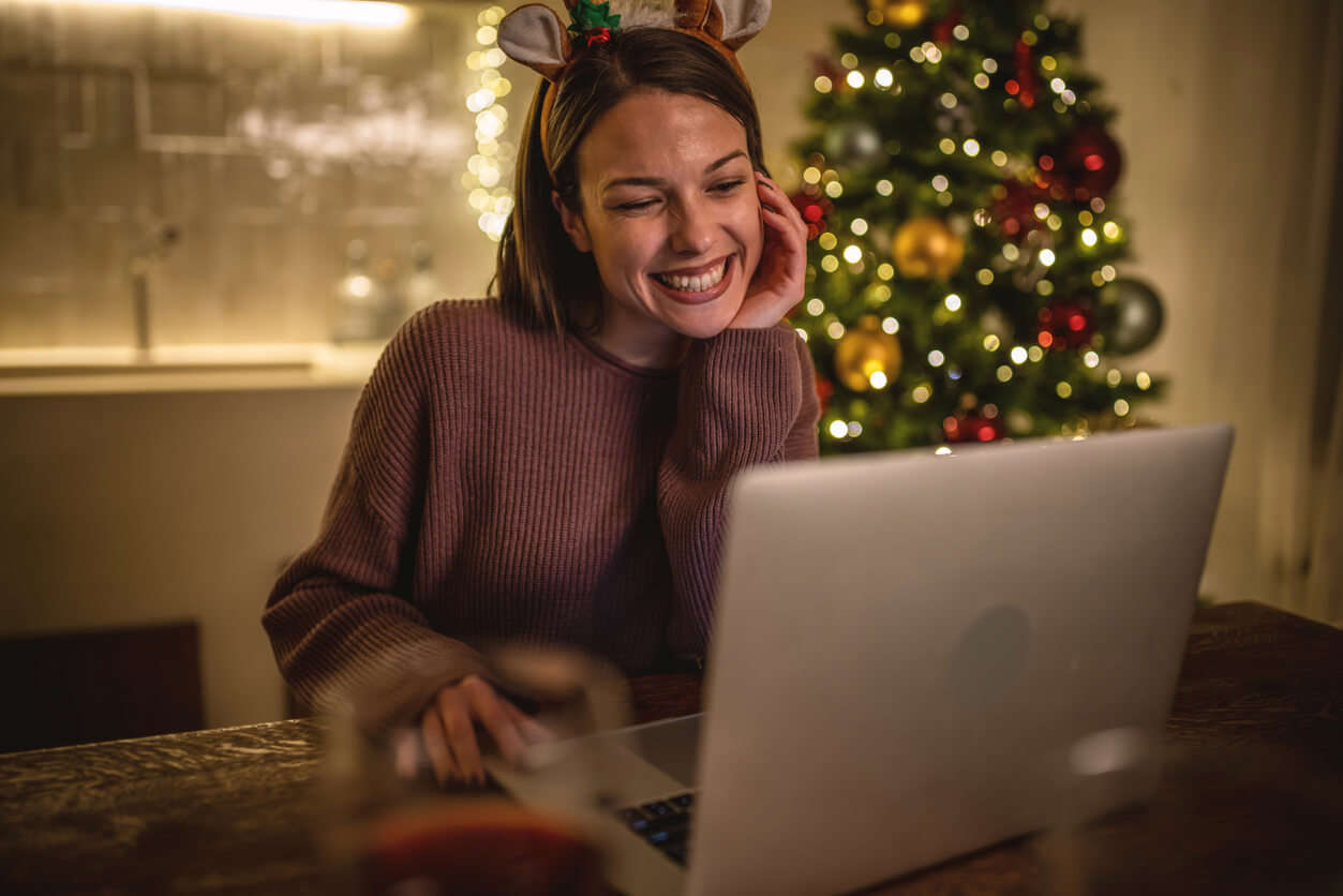 A lady wearing novelty Christmas reindeer ears sat at a table smiling whilst on a virtual call using her laptop with a Christmas tree in the foreground