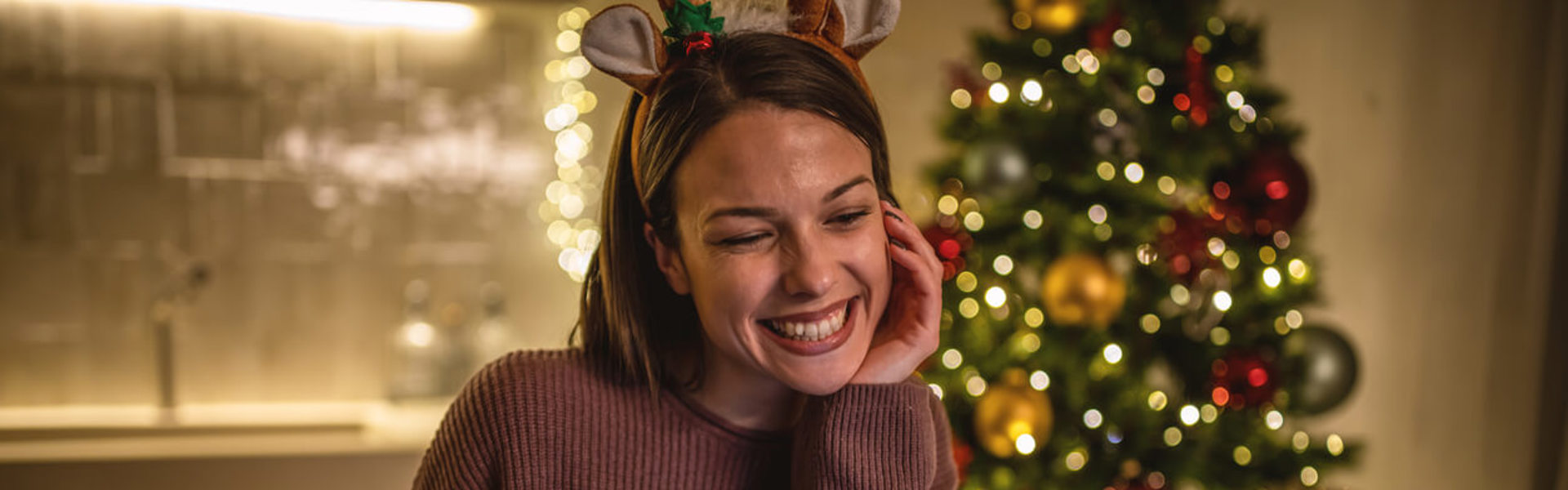 A lady wearing novelty Christmas reindeer ears sat at a table smiling whilst on a virtual call using her laptop with a Christmas tree in the foreground