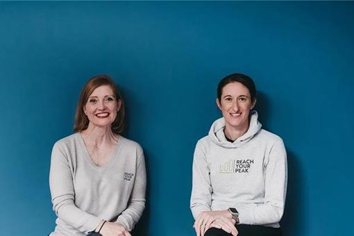 A photo of Reach You Peak Physiotherapists Sally and Maria, sat with their hands in their laps, smiling in front of a blue backdrop.
