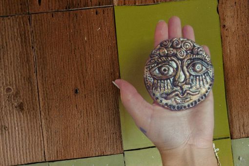 Main image: an ancient face medallion made out of air dry clay, a Greek-inspired art style, coloured with metallic pens/markers.