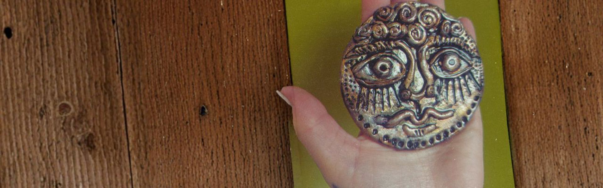 Main image: an ancient face medallion made out of air dry clay, a Greek-inspired art style, coloured with metallic pens/markers.