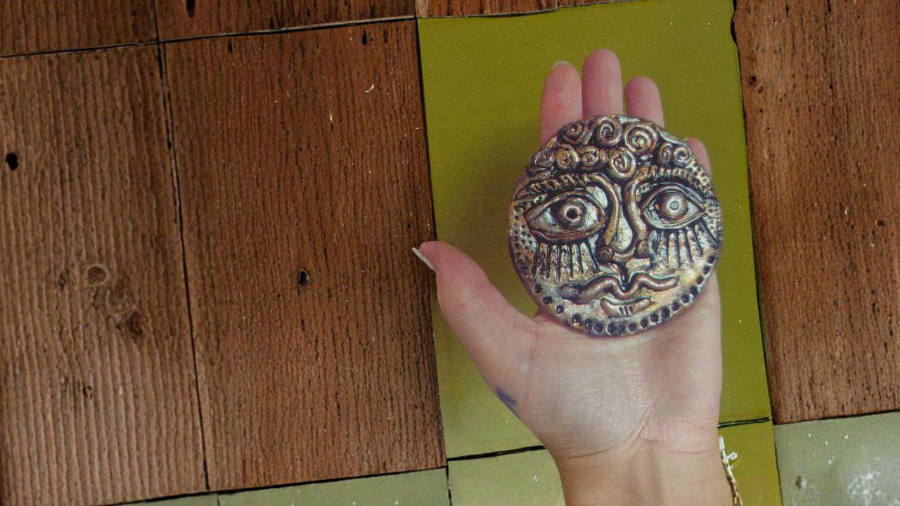 Main image: an ancient face medallion made out of air dry clay, a Greek-inspired art style, coloured with metallic pens/markers.
