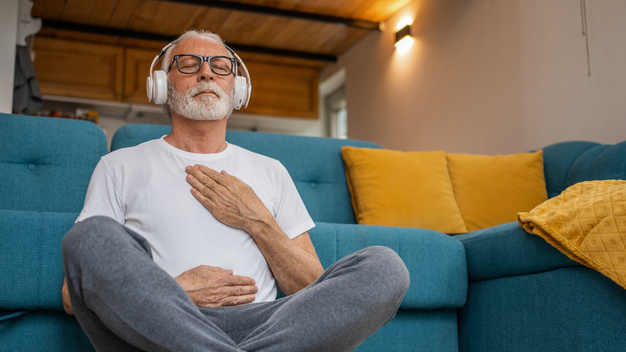 Main Image: a man meditating in his lounge, wearing headphones with his legs crossed and a hand on his chest.