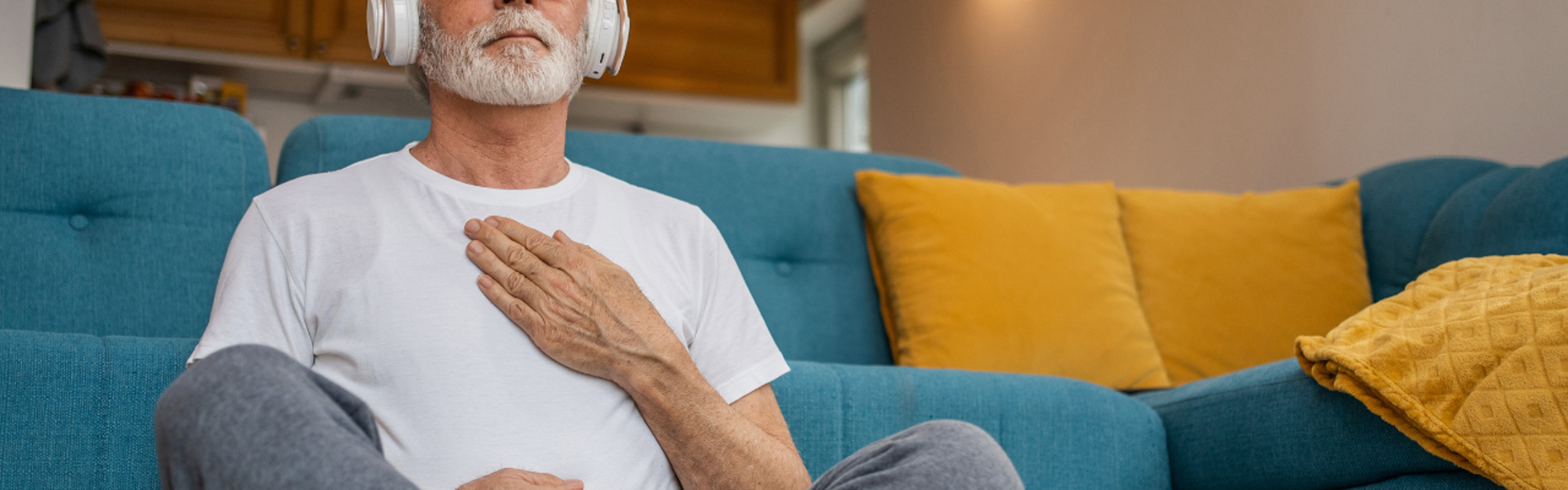 Main Image: a man meditating in his lounge, wearing headphones with his legs crossed and a hand on his chest.