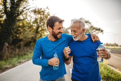 A young man with brown hair and a dark bear and an older man with white hair and a white beard wearing blue exercise tops arm in arm and celebrating