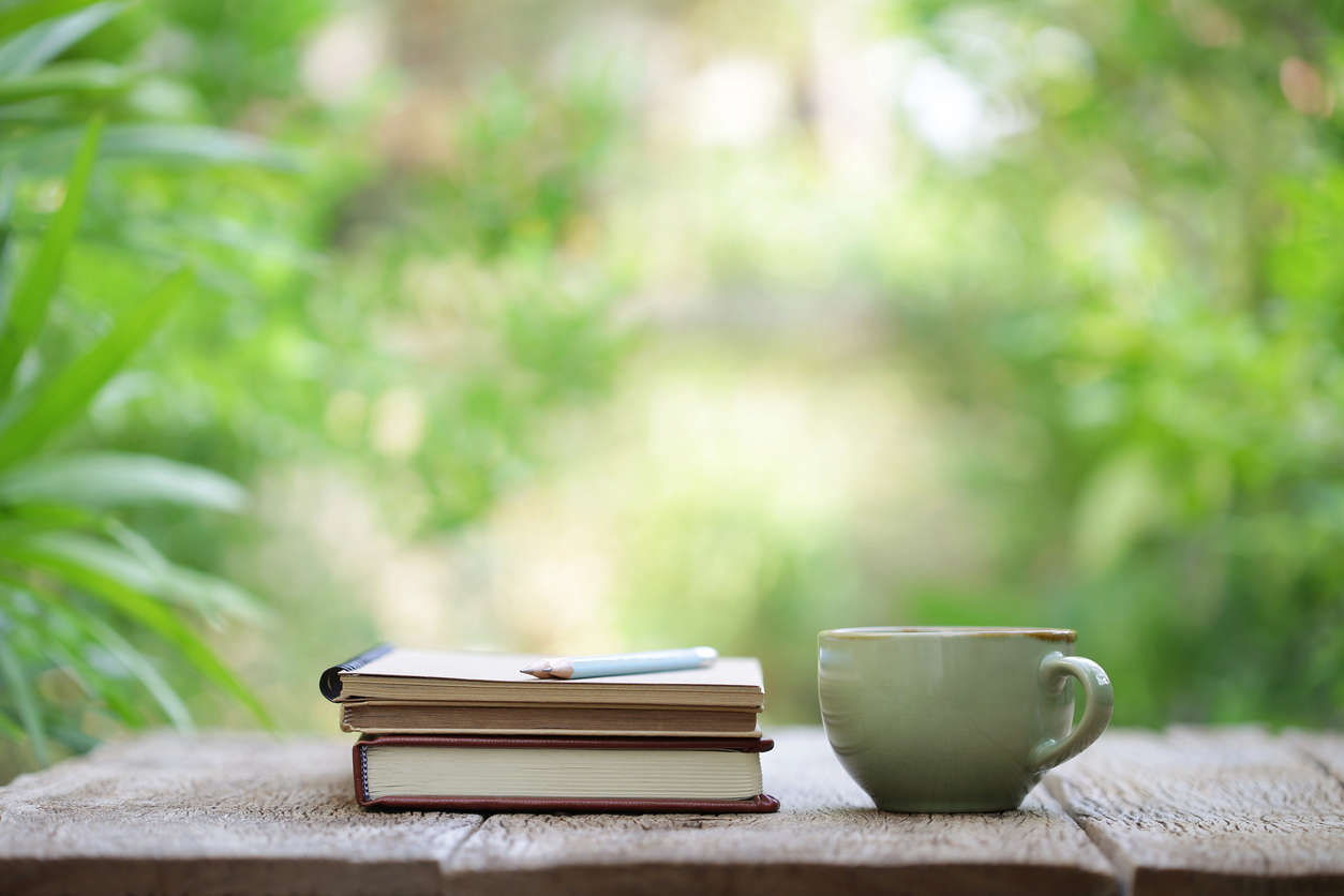 a pile of writing books with a blue pencil resting on top on a wooden table with a green outdoor background