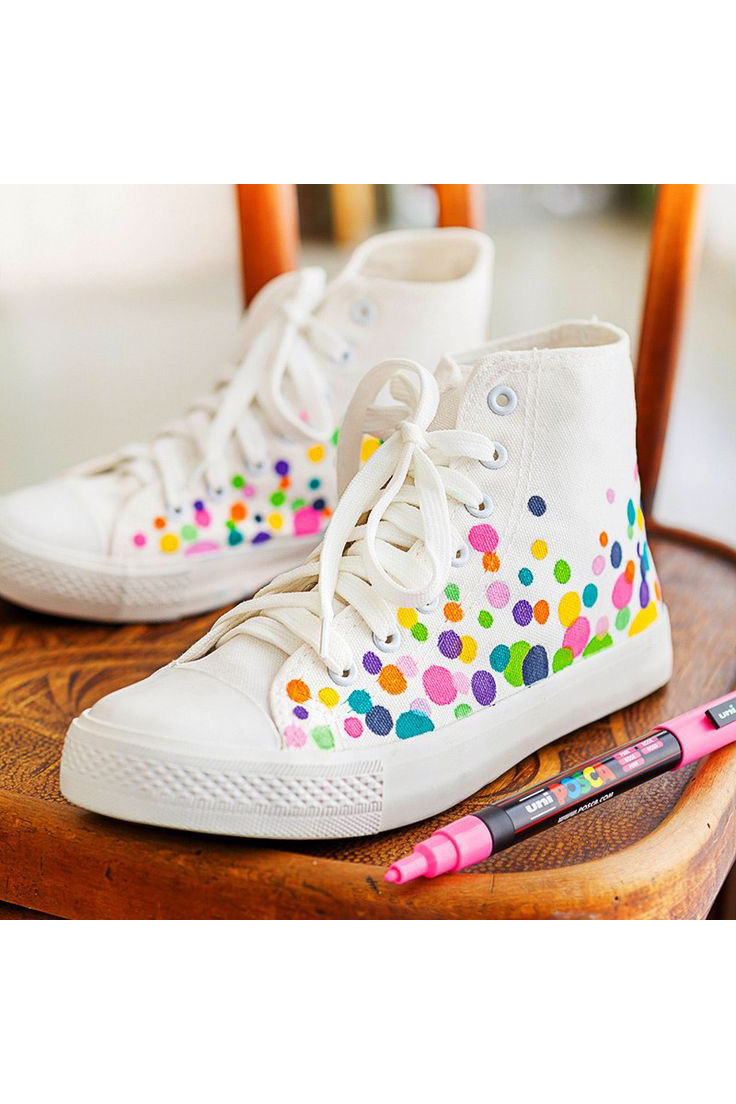 A pair of white canvas shoes / trainers with a colourful fabric marker polka dot design, with a pink fabric POSCA marker next to them on a wooden chair
