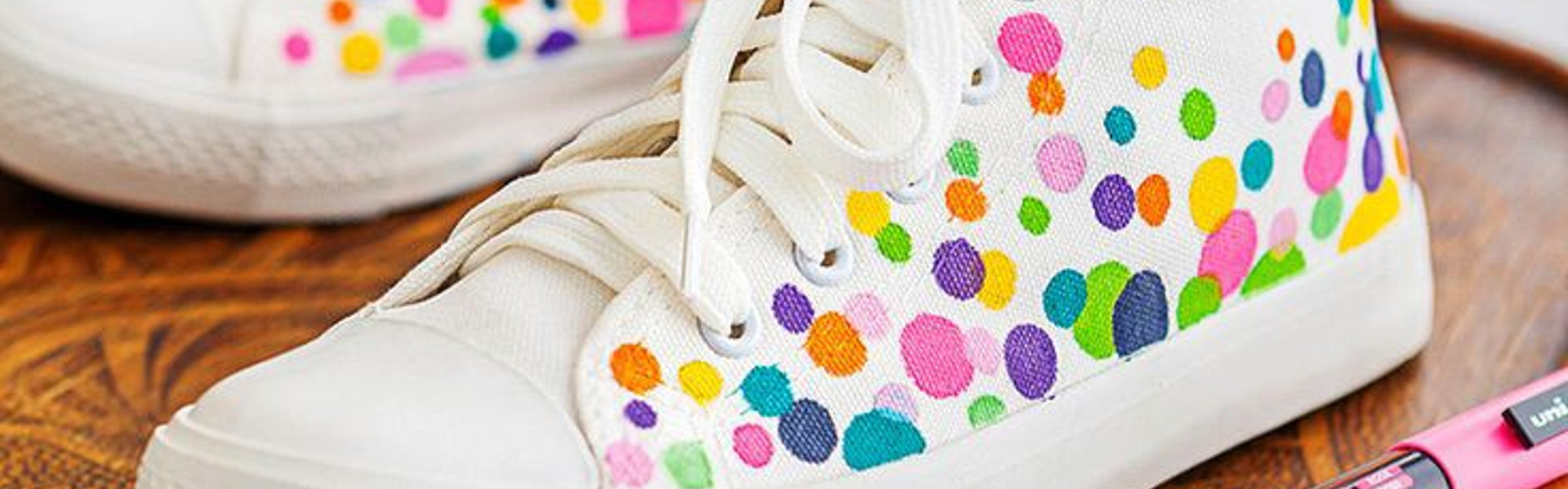 A pair of white canvas shoes / trainers with a colourful fabric marker polka dot design, with a pink fabric POSCA marker next to them on a wooden chair