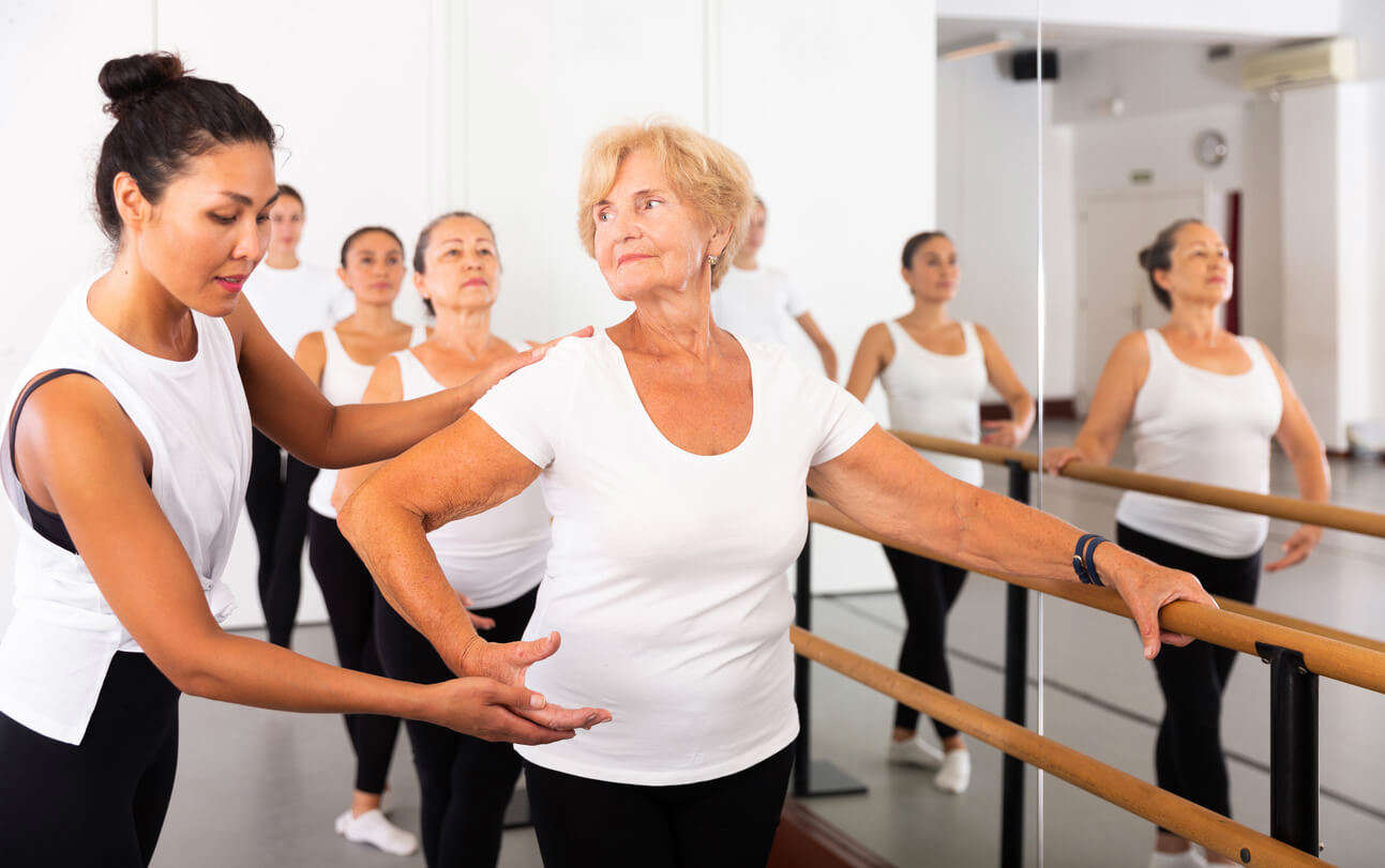 An elderly lady doing ballet in a ballet studio holding onto the bar with support from another woman