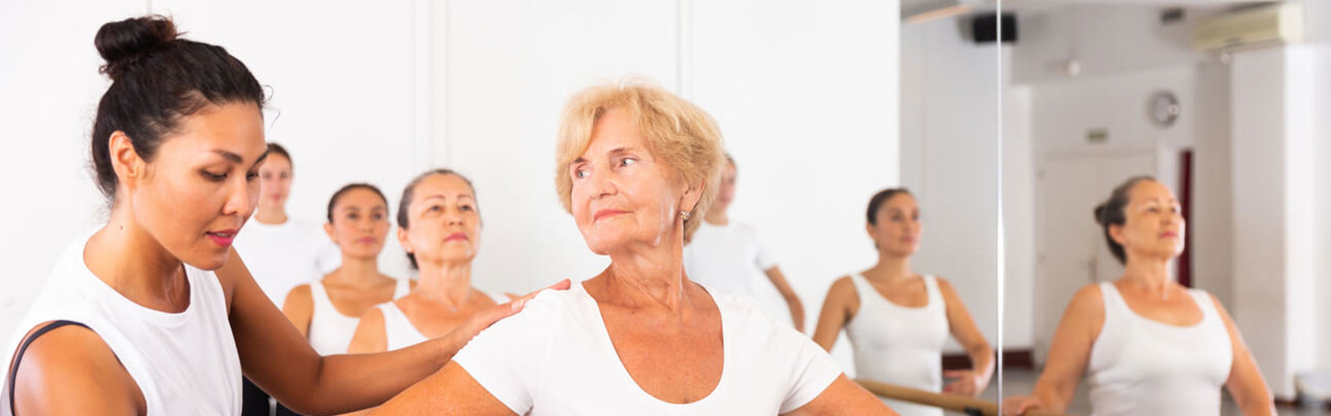 An elderly lady doing ballet in a ballet studio holding onto the bar with support from another woman