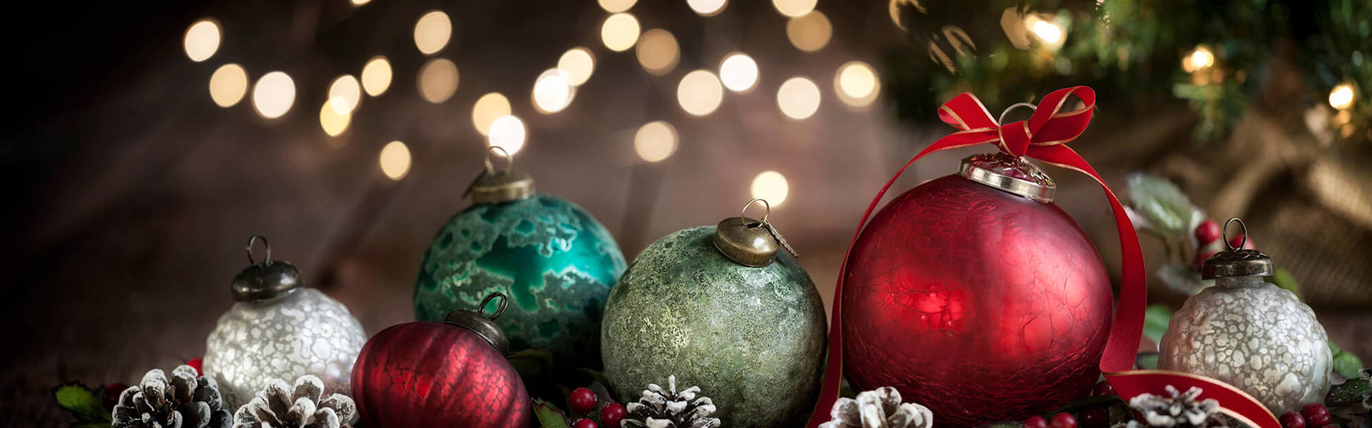 A row of red, green and gold Christmas tree decoration baubles lined up surrounded by red velvet ribbon and snow frosted pinecones