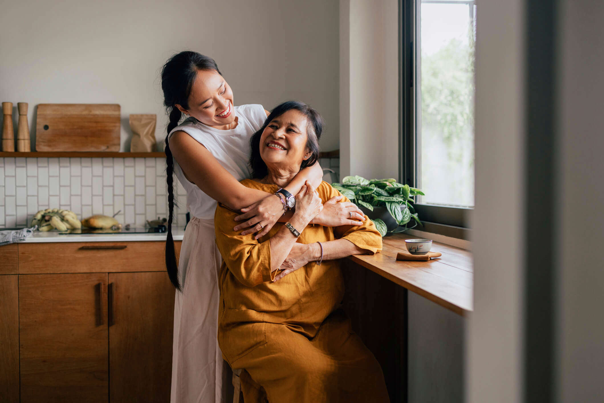 A Happy Woman Hugging Her Mother While She Is Sitting In The Kitchen And Drinking Tea