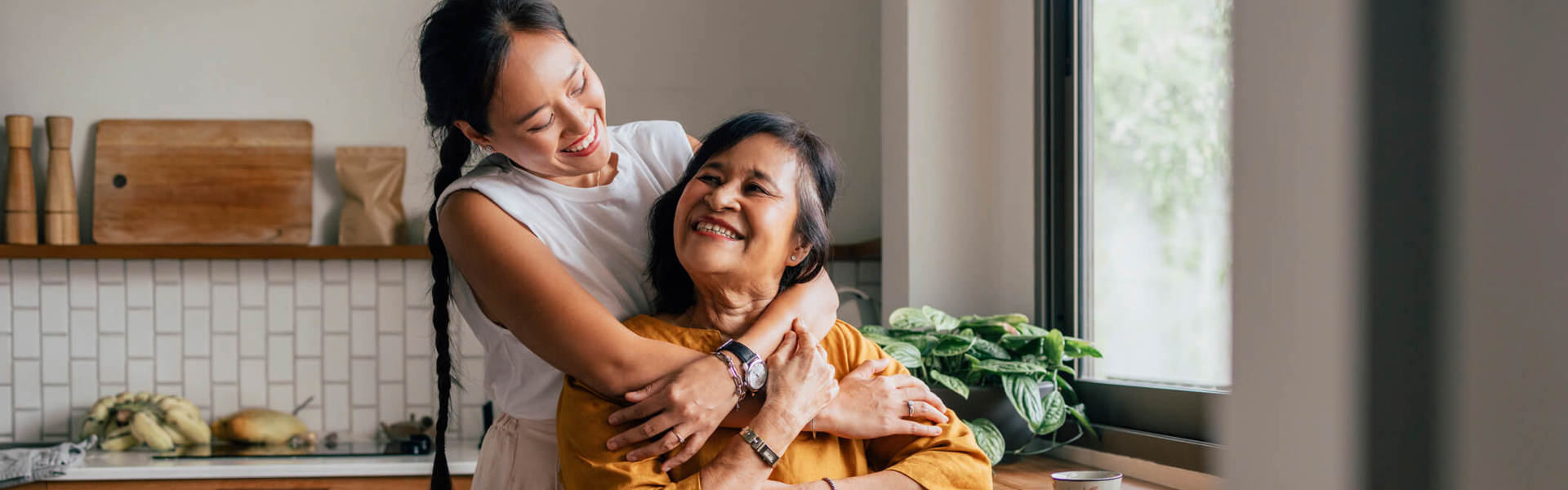 A Happy Woman Hugging Her Mother While She Is Sitting In The Kitchen And Drinking Tea
