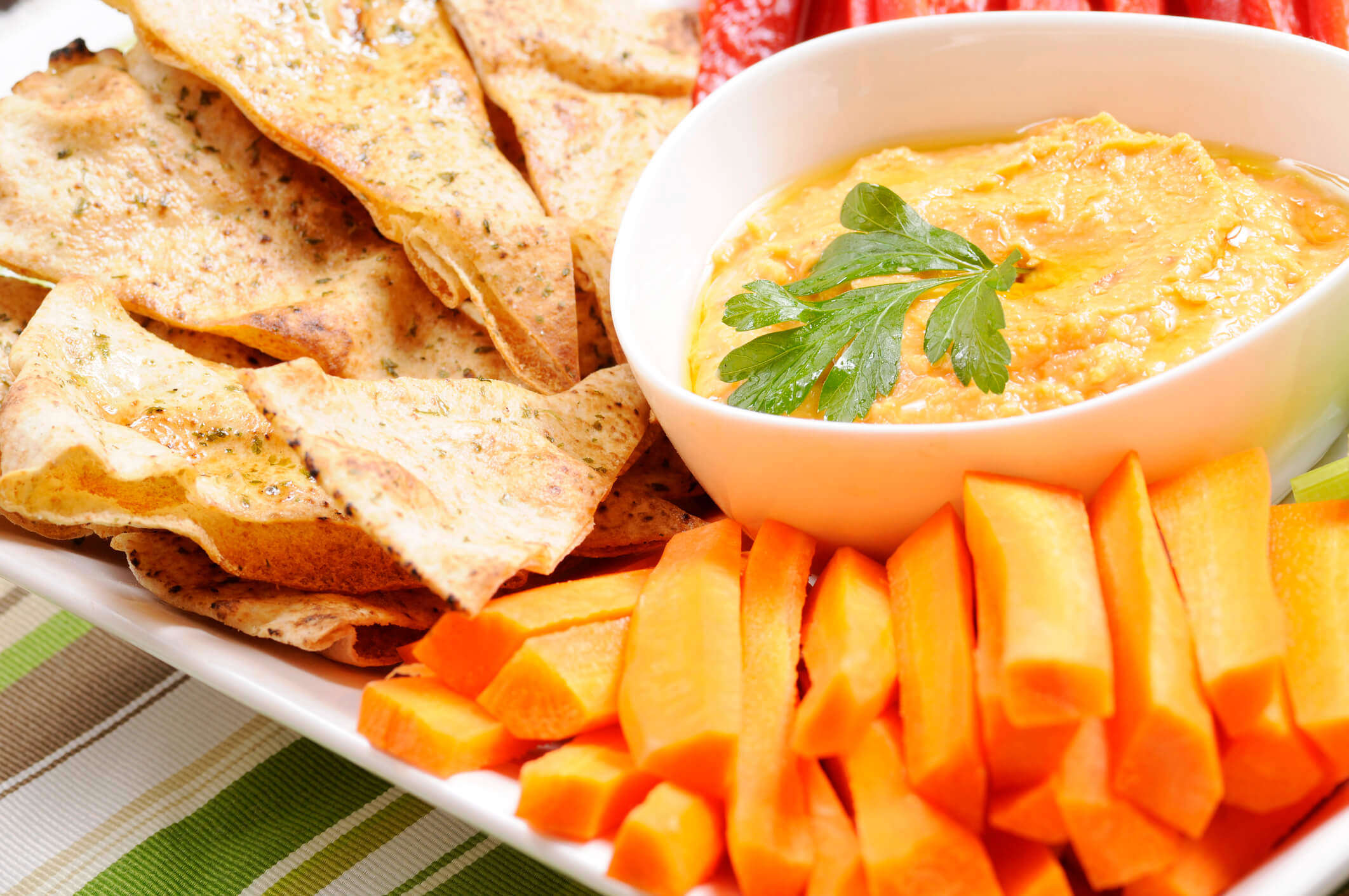 homemade hummus in a white bowl surrounded by carrot sticks and tortilla chips