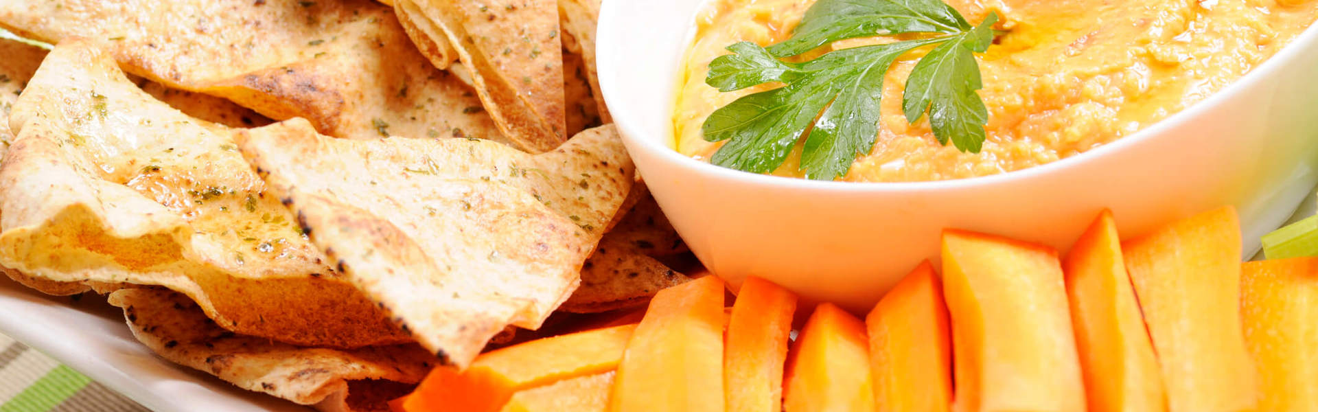 homemade hummus in a white bowl surrounded by carrot sticks and tortilla chips