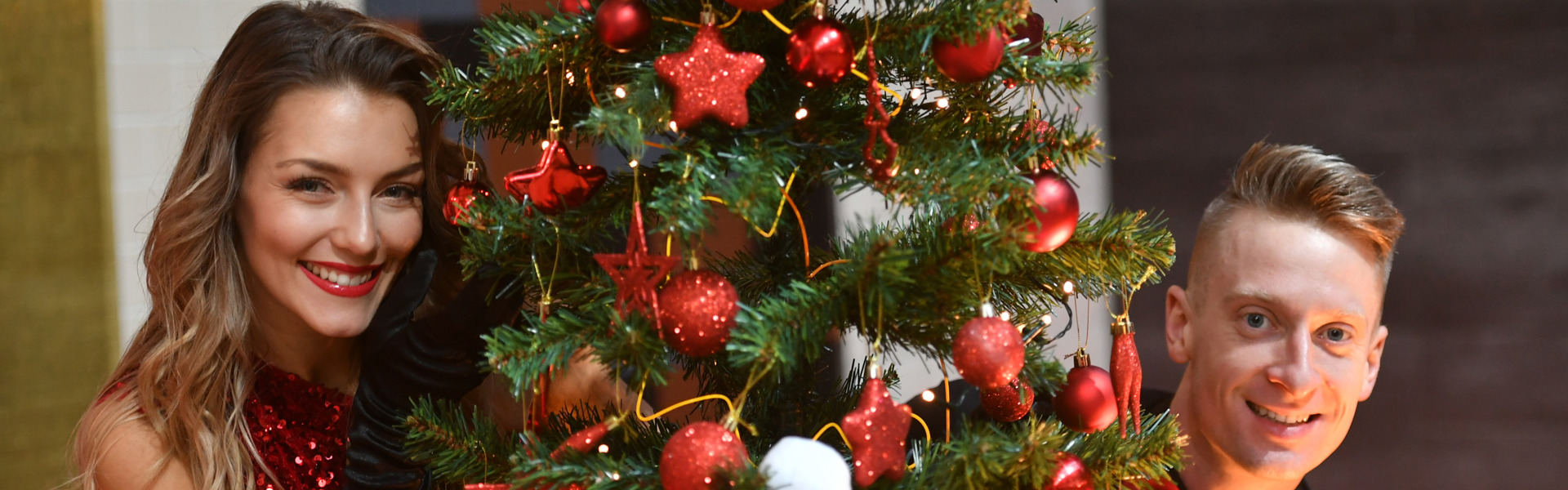 Ballroom dancers Michael Danilczuk and Jowita Pryztal standing either side of a decorated Christmas tree and smiling