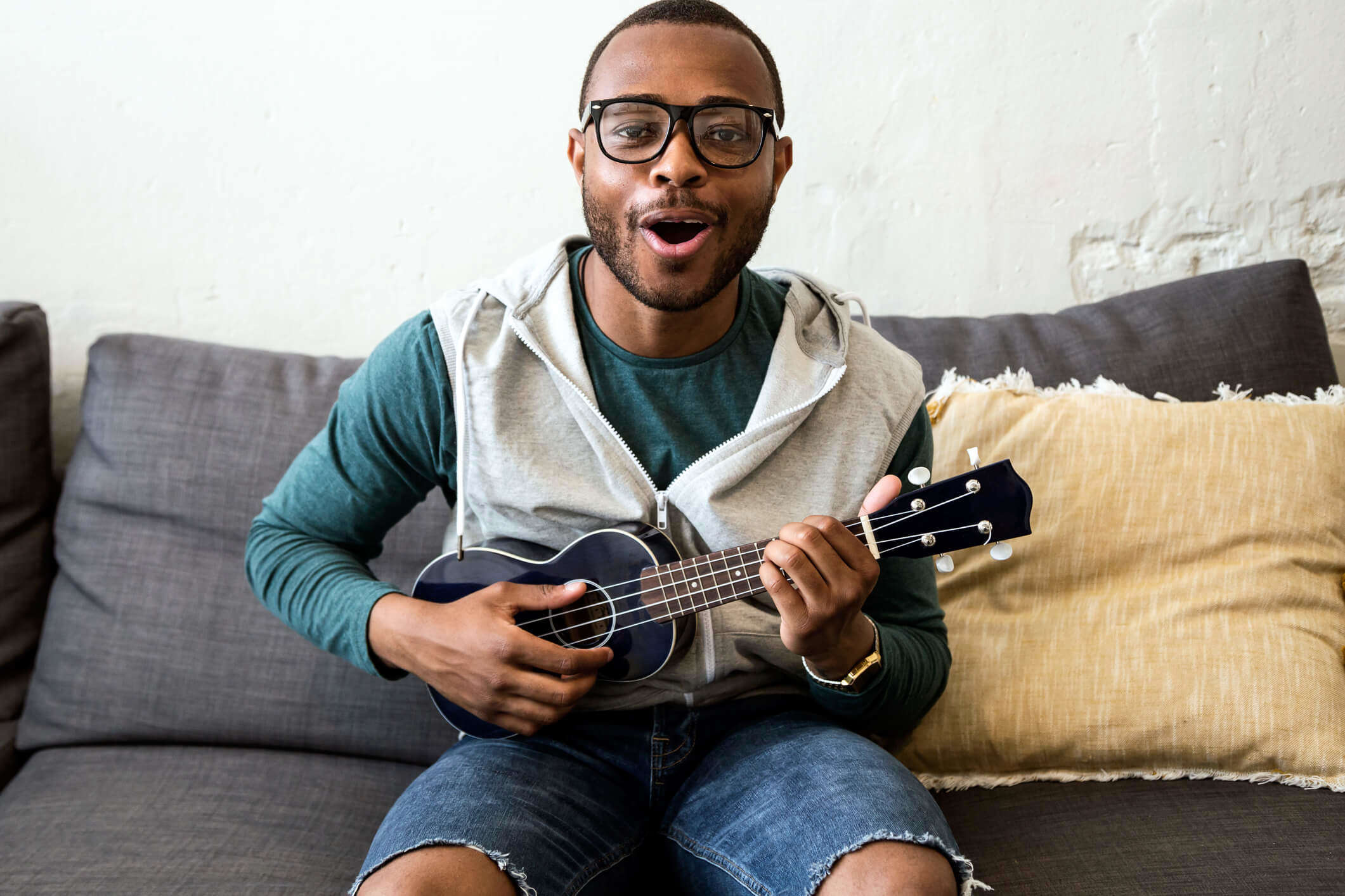 a man wearing glasses sat on the sofa playing the ukulele and singing 