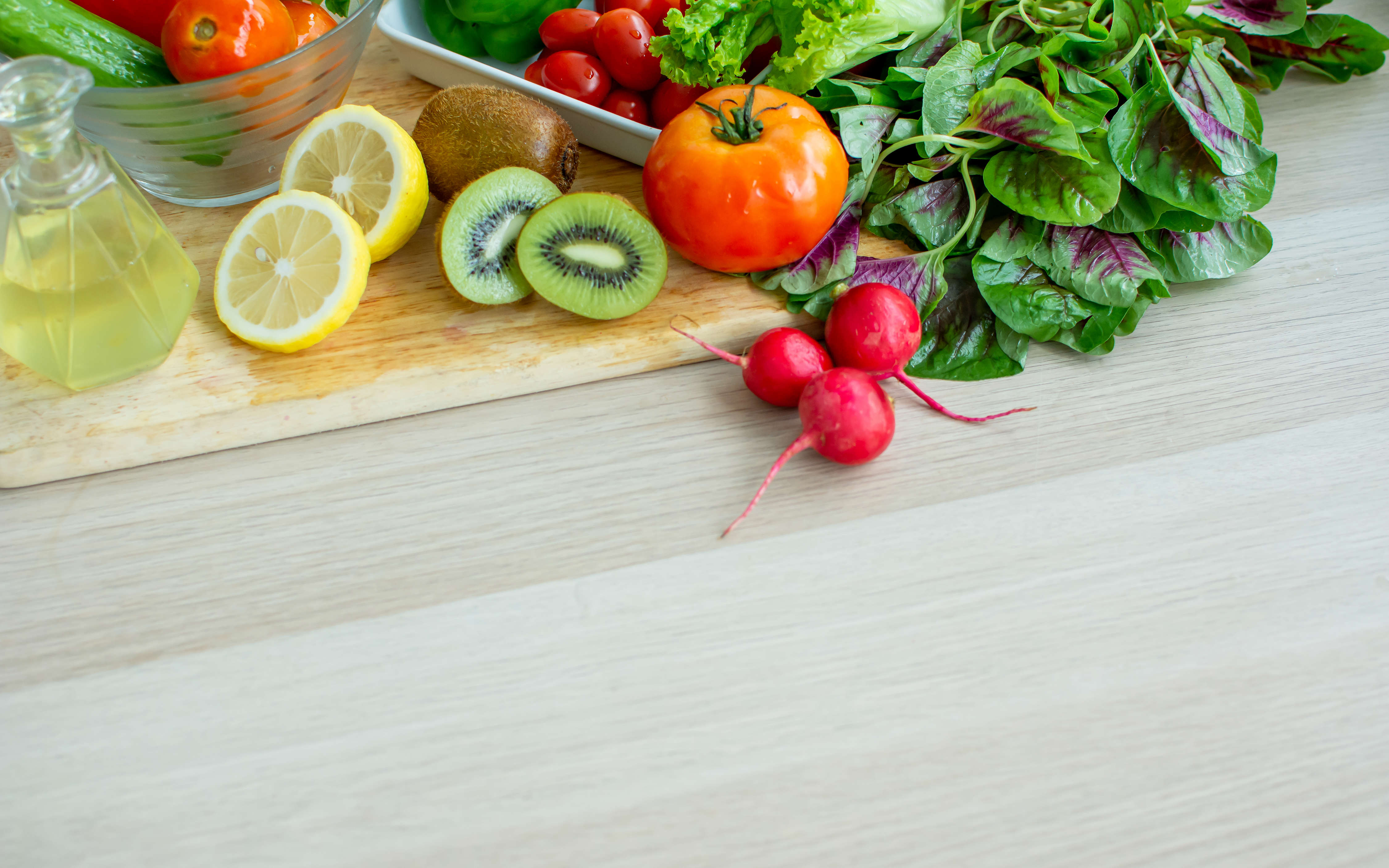 Main Image; An assortment of fruits and vegetables on a wooden chopping board including, lemons, kiwis, radishes, tomatoes and salad leaves.