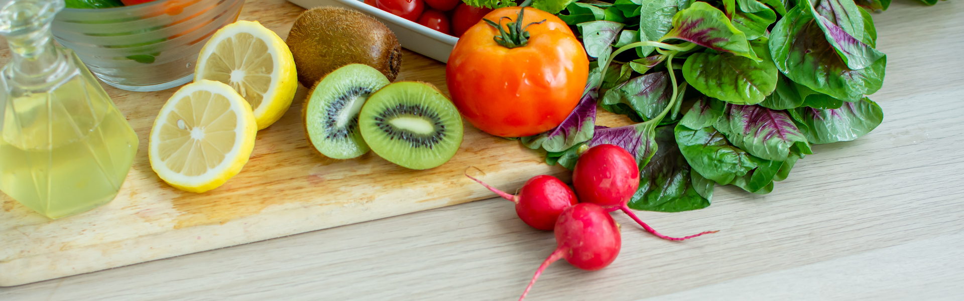 Main Image; An assortment of fruits and vegetables on a wooden chopping board including, lemons, kiwis, radishes, tomatoes and salad leaves.