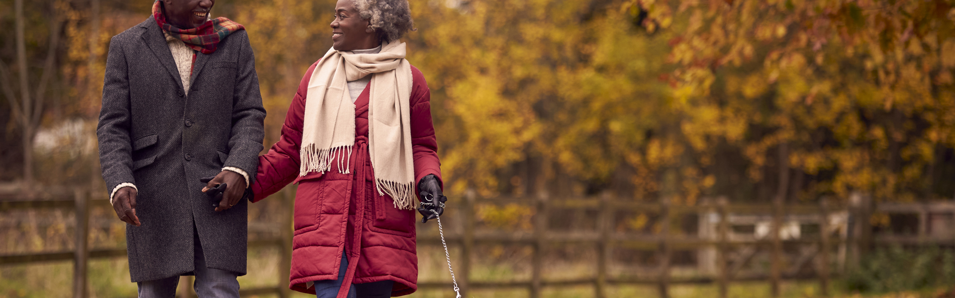 Main image: an older couple walking their dog in the park, in autumn with orange leaves in the trees and scattered around the path.