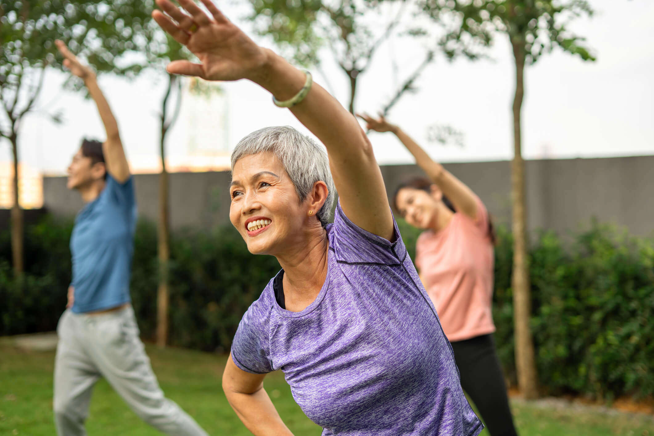 Woman in a purple workout t-shirt stretching her left arm over her head, with her other hand on her hip, exercising outdoors with two people also doing the same stretch behind her.