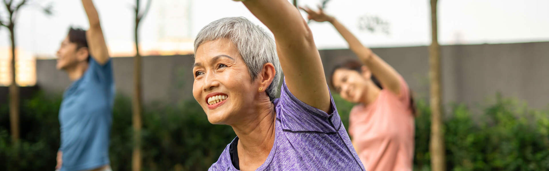 Woman in a purple workout t-shirt stretching her left arm over her head, with her other hand on her hip, exercising outdoors with two people also doing the same stretch behind her.