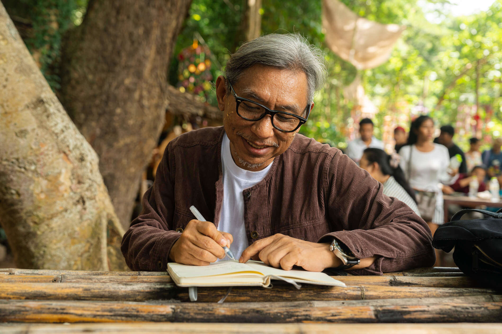 A smiling man enjoying a peaceful moment sat journaling at a wooden table outside under a tree.