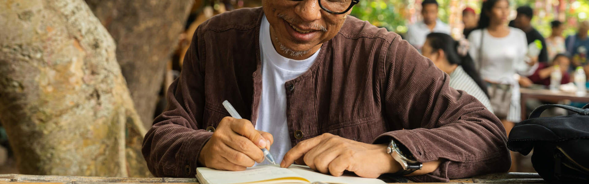 A smiling man enjoying a peaceful moment sat journaling at a wooden table outside under a tree.