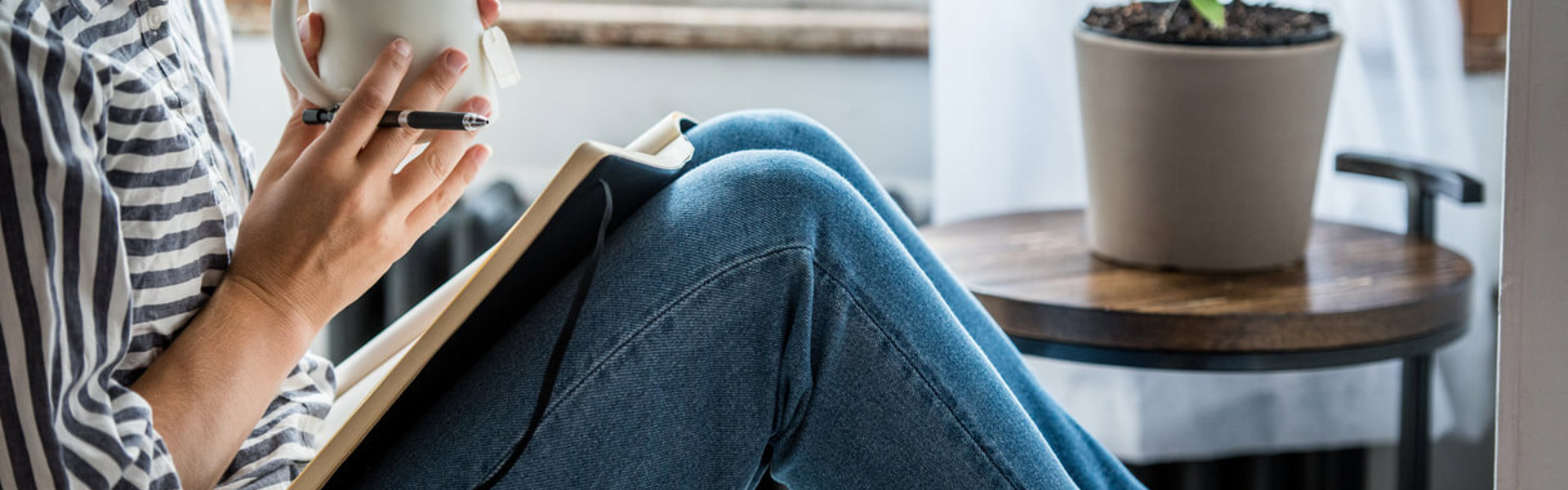 a person wearing a stripe shirt and jeans sat holding a mug of tea with a pen and open book on their lap