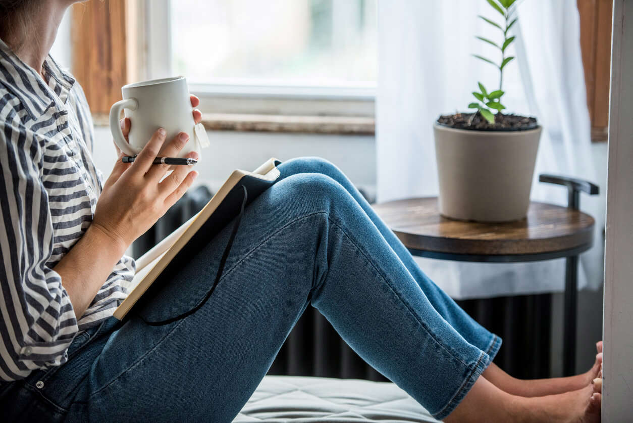 a person wearing a stripe shirt and jeans sat holding a mug of tea with a pen and open book on their lap