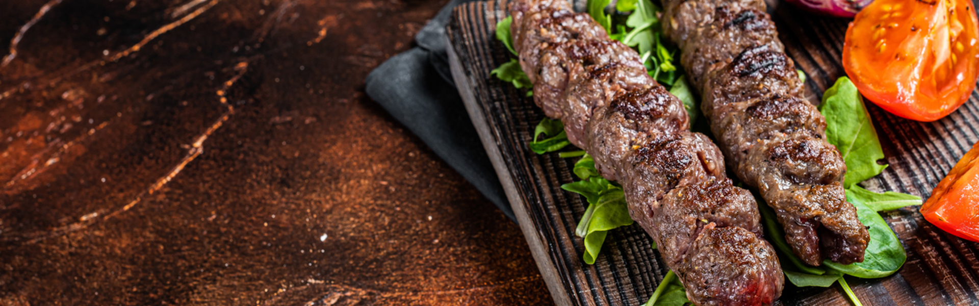 Two grilled meat koftas on wooden board with tomatoes and herbs