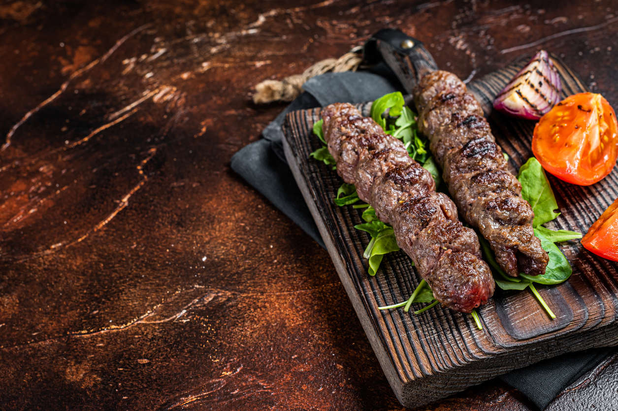 Two grilled meat koftas on wooden board with tomatoes and herbs