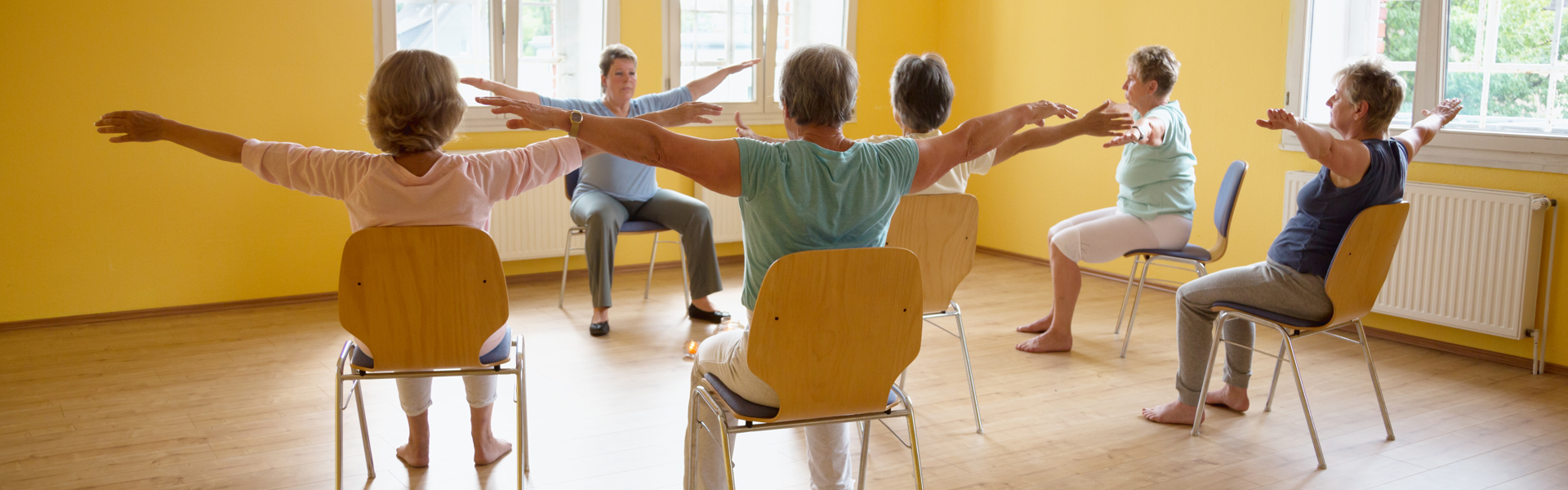 Main image: a group of people taking part in a seated Parkinson's dance class, in a bright yellow room.