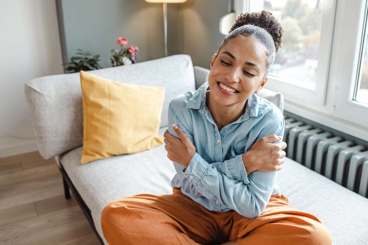 a woman wearing a denim shirt and orange trousers sat cross legged on a sofa giving herself a hug