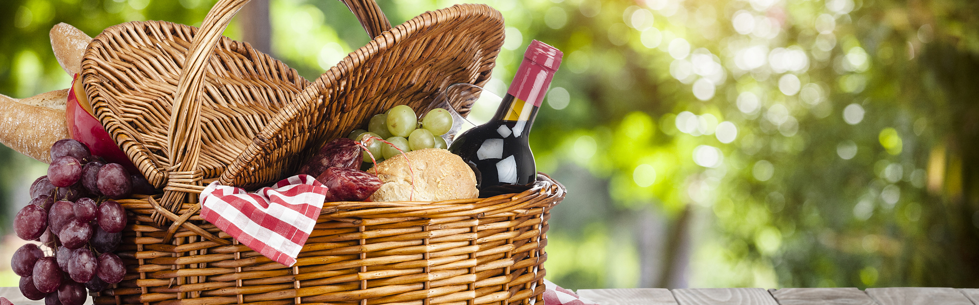 a picnic basket on a red and white check gingham pattern picnic blanket filled with wine and cheese and grapes