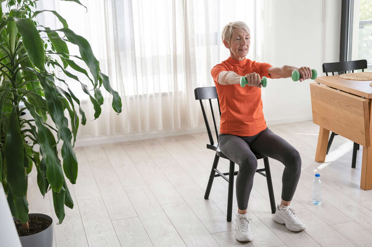 Smiling beautiful senior woman health instructor doing chair exercises with dumbbells