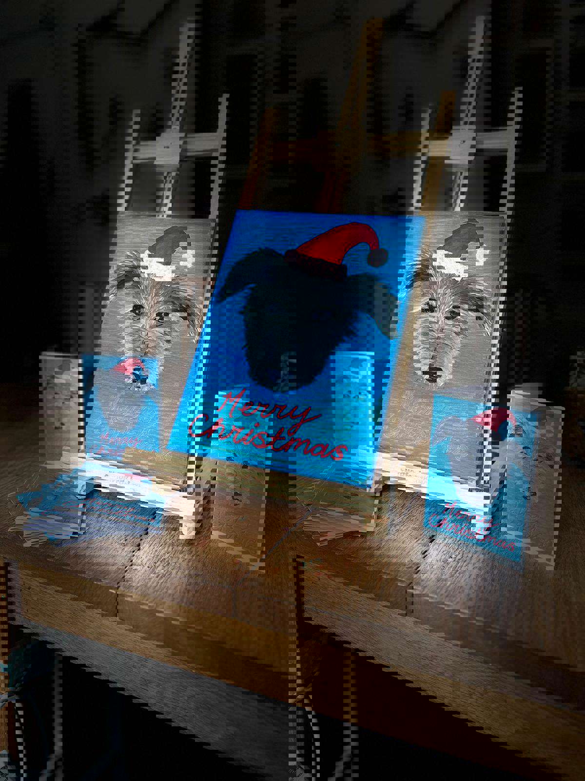 an easel with a portrait of a pet dog wearing a Christmas hat on a blue back ground