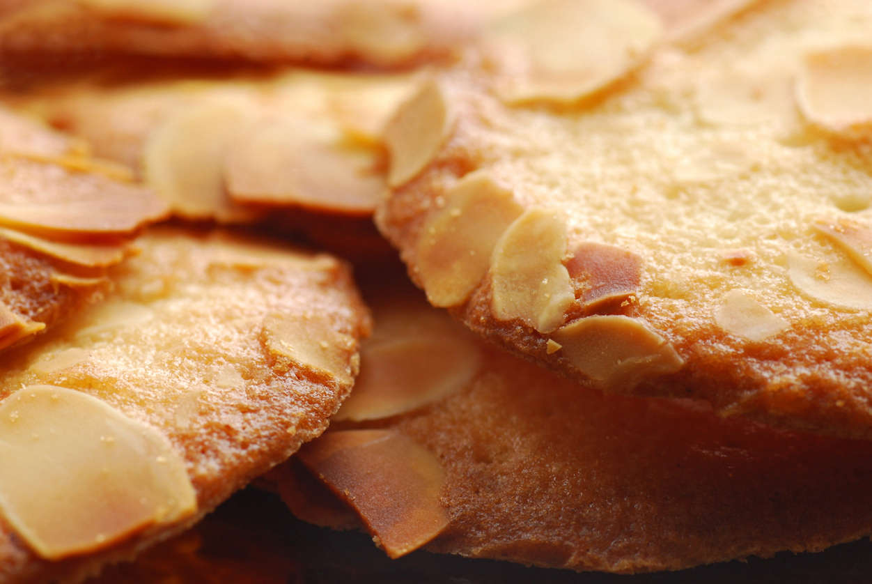 Close-up of several thin, crispy almond cookies, each topped with sliced almonds and a golden-brown surface.