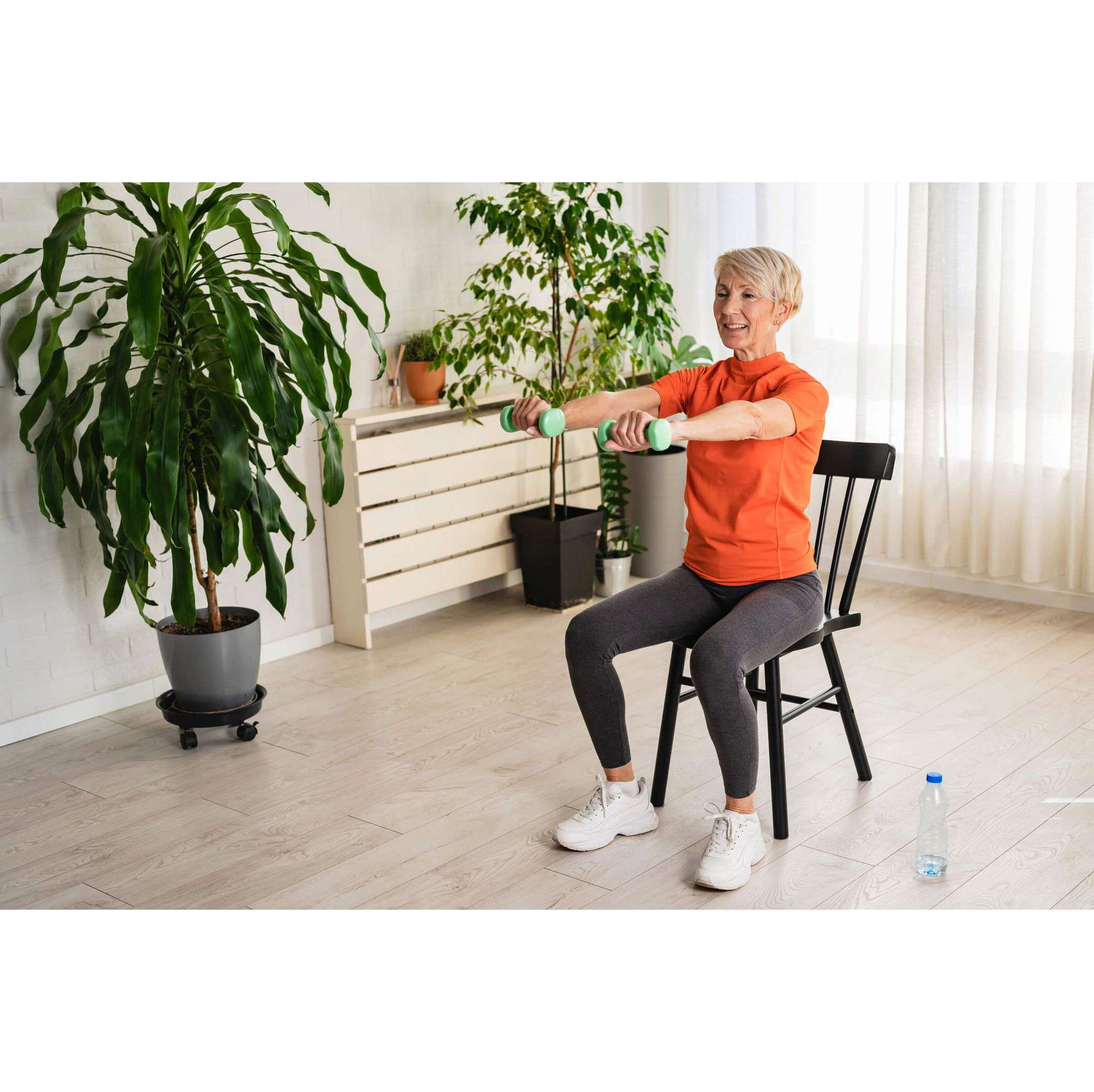 A woman sat on a chair, holding two small green dumbbells out in front of her, wearing a bright orange top and smiling. With a variety of plants in the background.