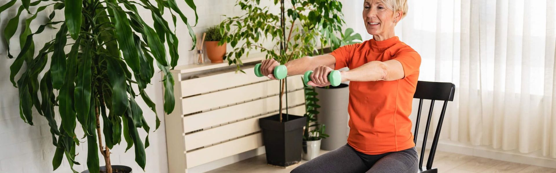 A woman sat on a chair, holding two small green dumbbells out in front of her, wearing a bright orange top and smiling. With a variety of plants in the background.