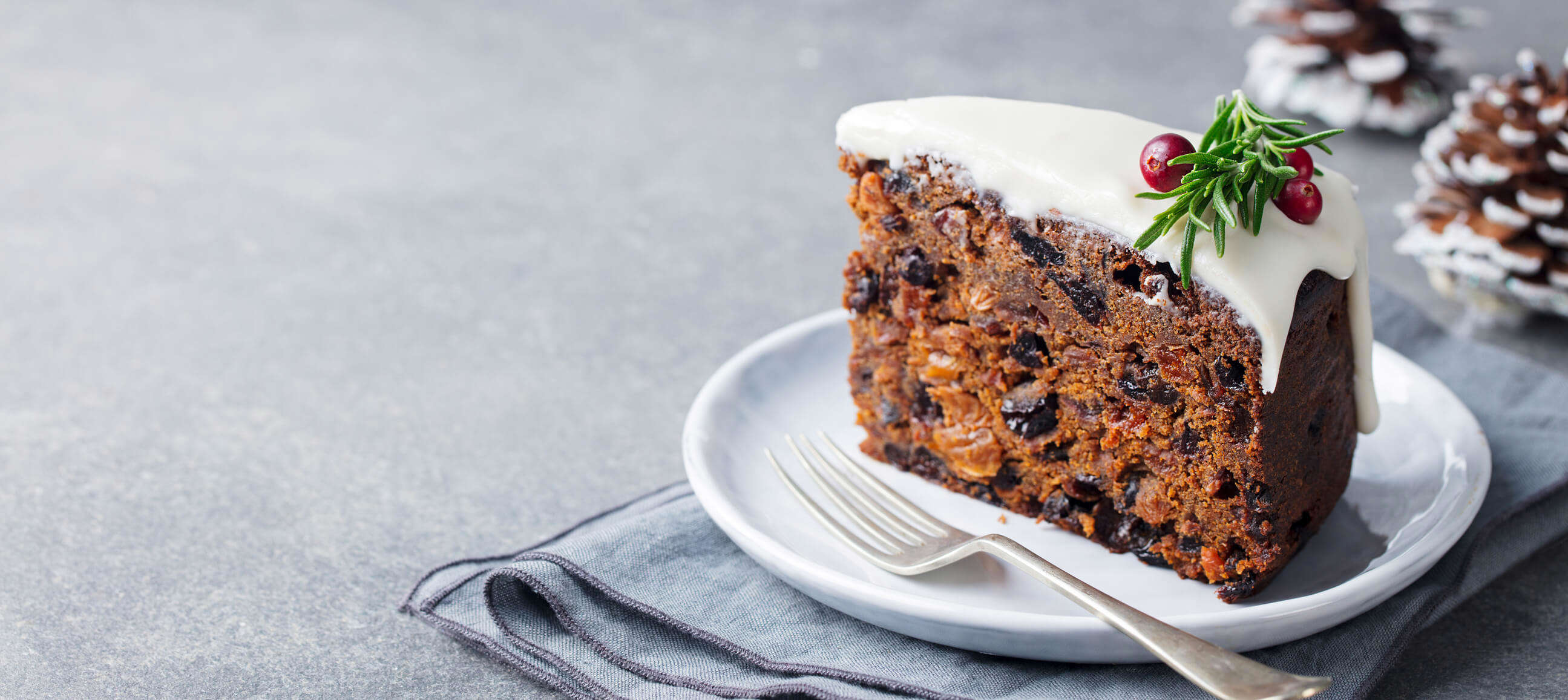 A slice of Christmas cake on a white plate with a small fork next to it and decorating with red berries and rosemary