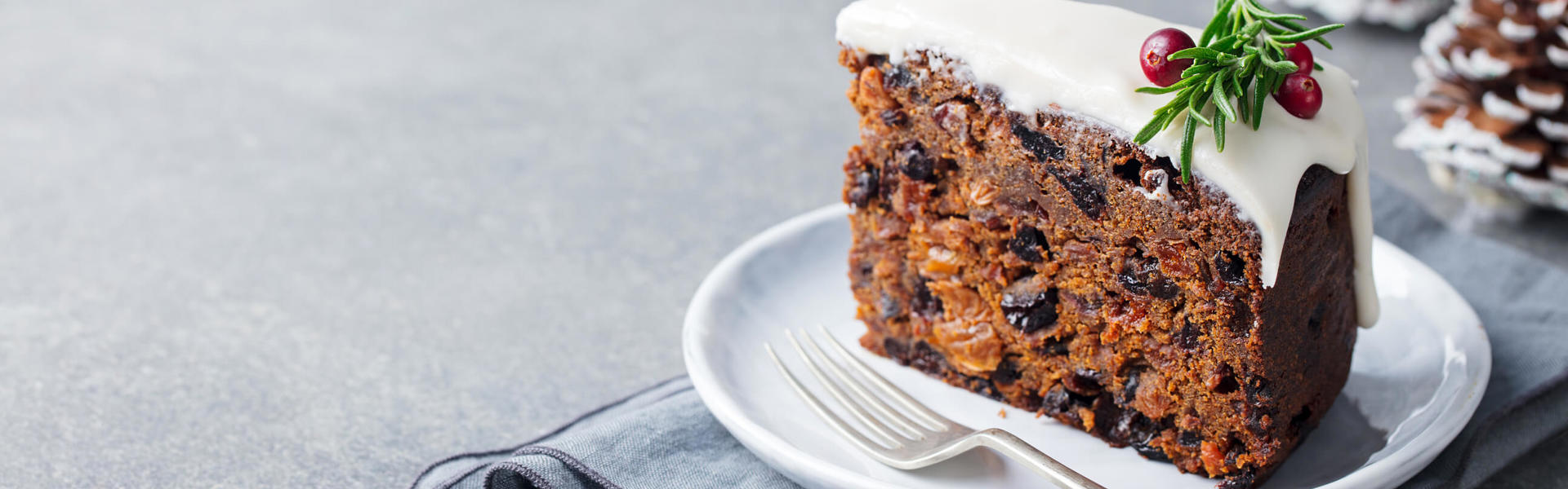 A slice of Christmas cake on a white plate with a small fork next to it and decorating with red berries and rosemary
